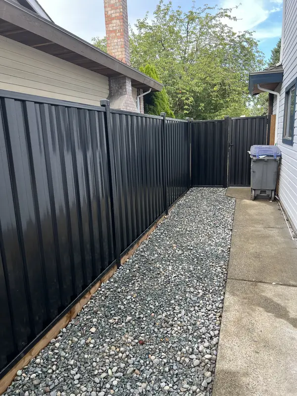 Black steel privacy fence and gate on a timber base, with river rock along a side yard