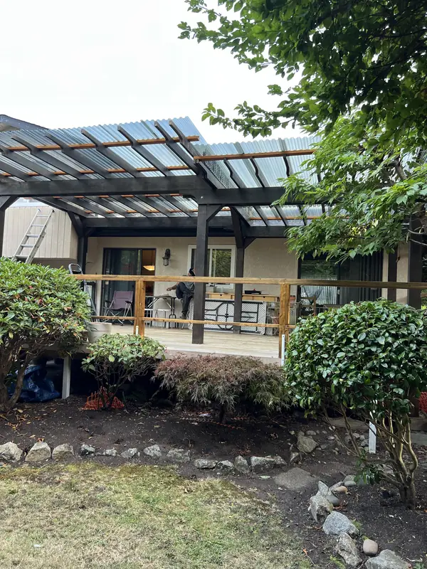 Pergola with clear roof panels over a back patio, with a rock border and shrubs below