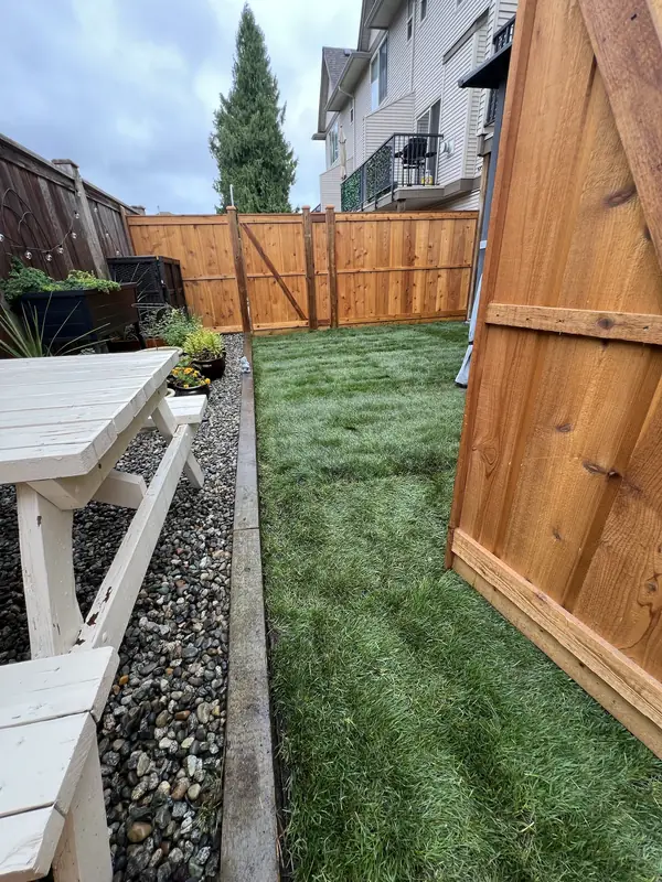 New sod lawn in a narrow backyard, with a river rock strip and timber edging on one side and a cedar fence at the back