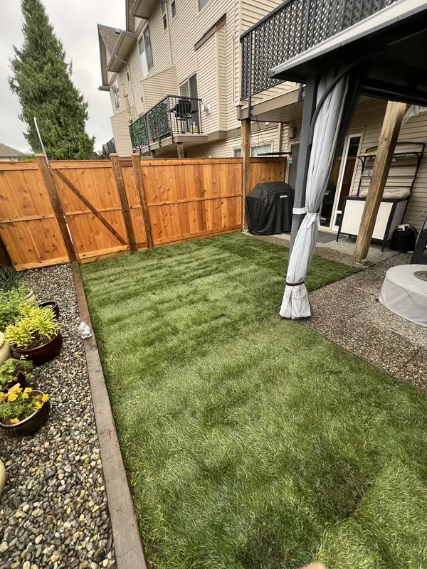 New sod lawn in a townhouse backyard beside a stained cedar fence and gate, with a river rock border